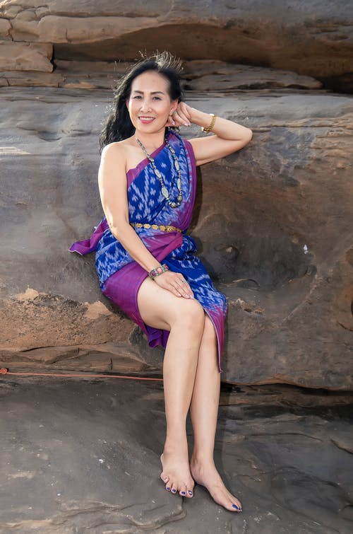 微笑的女人，穿着蓝色和紫色的花无袖连衣裙，坐在棕色岩石上的照片 · 免费素材图片