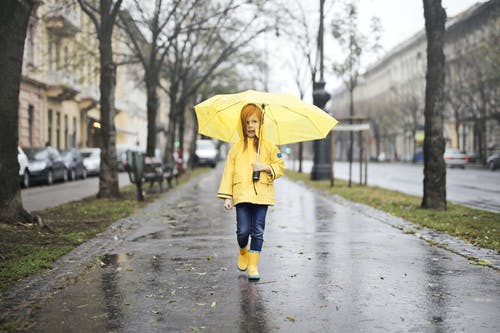 黄色雨衣和黄色靴子的女孩拿着黄色的雨伞，在灰色的混凝土人行道上行走 · 免费素材图片