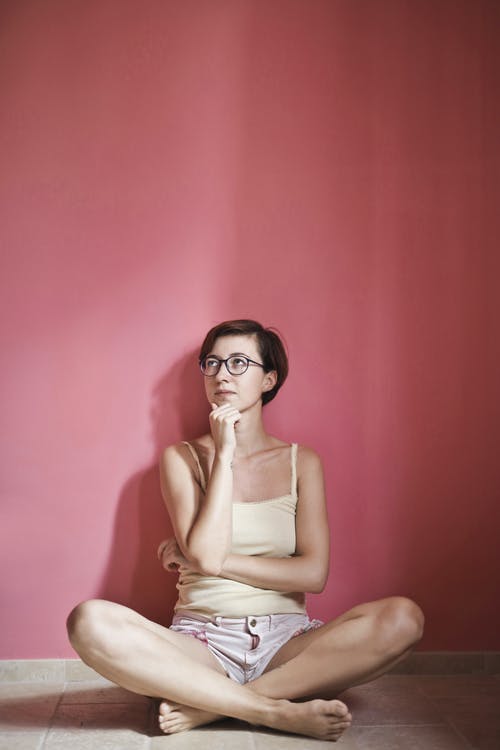 黄色背心和粉红色短裤坐在粉红色的墙壁旁边的地板上有思想的女人的照片 · 免费素材图片