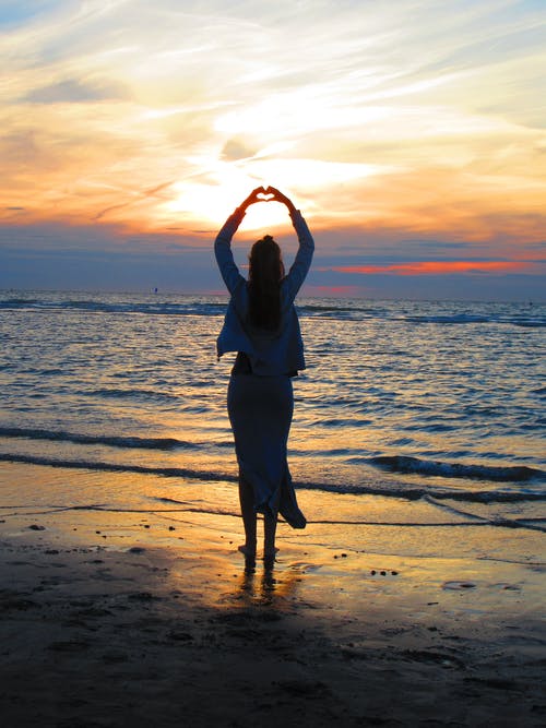 女人，双臂高举使心脏标志，而站在海滩上在日落时 · 免费素材图片