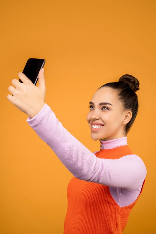 微笑的女人站在橙色背景前的同时采取自己的自拍照的照片 · 免费素材图片