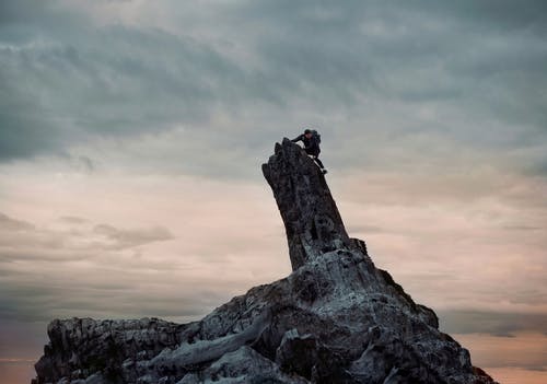穿黑夹克的男人爬上多云的天空下的岩层 · 免费素材图片