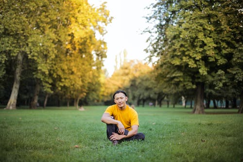 穿着橙色t恤的人的照片，同时拿着他的智能手机，独自坐在绿草地上 · 免费素材图片