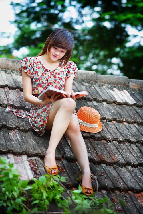 女人穿着白色，绿色和红色的花帽袖子的裙子在绿树附近的黑色屋顶上读书 · 免费素材图片