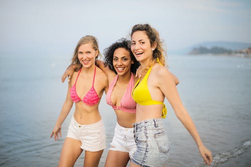 在比基尼泳装上衣和牛仔短裤站在海滩上的三个微笑妇女的照片 · 免费素材图片