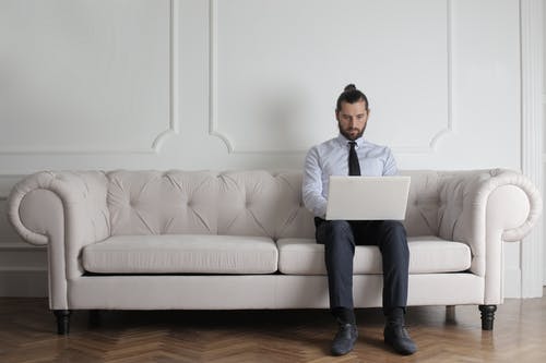 身穿蓝色上衣和领带坐在白色沙发上使用笔记本电脑的人 · 免费素材图片