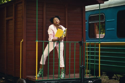 女人穿着粉红色西装外套，裤子和绿色的高跟鞋，拿着黄色的盖的书 · 免费素材图片
