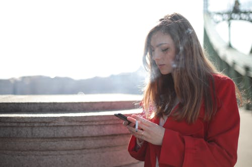 拿着智能手机吸烟的红色外套的女人 · 免费素材图片