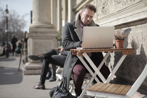 穿黑夹克的男人坐在使用macbook的棕色木制折叠椅上 · 免费素材图片
