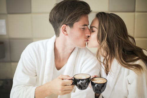 夫妇在白色浴袍，拿着黑色的陶瓷咖啡时接吻的照片 · 免费素材图片