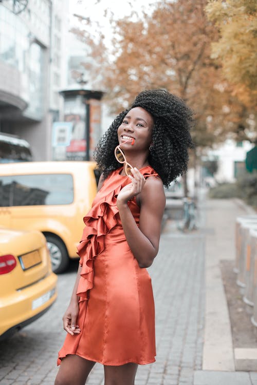 微笑的女人，穿着橙色无袖连衣裙站在人行道上，同时拿着眼镜的照片 · 免费素材图片