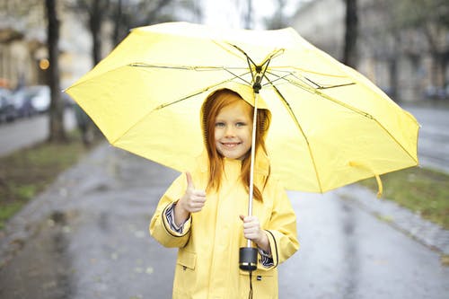 微笑的女孩，穿着黄色的雨衣，拿着一把黄色的雨伞，放弃一个大拇指的照片 · 免费素材图片