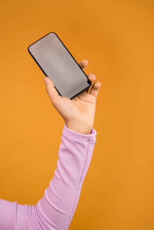 在橙色背景下手持黑色智能手机的人 · 免费素材图片