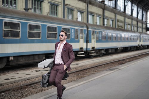 穿粉红色正装衬衫和棕色西装的男人背着行李走在火车旁边 · 免费素材图片