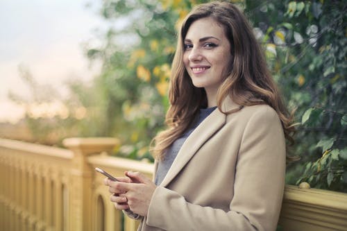 倾斜着石栏杆时拿着智能手机的棕色外套的微笑妇女的选择性聚焦照片 · 免费素材图片