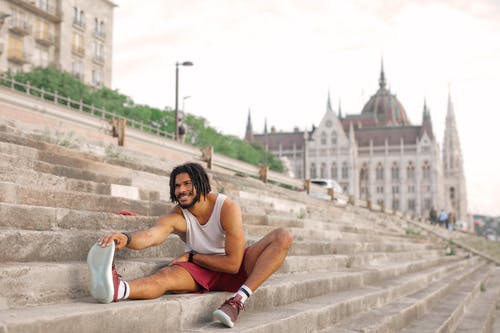 坐在混凝土楼梯上伸展的白色背心和红色短裤的男人微笑着的选择性焦点照片 · 免费素材图片