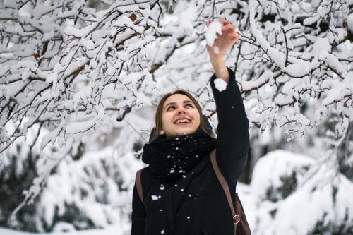 黑色的冬大衣，站在积雪的树下微笑的女人的照片 · 免费素材图片