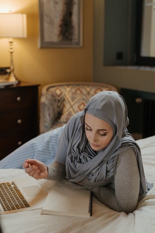 灰色盖头的女人在使用笔记本电脑时记笔记 · 免费素材图片
