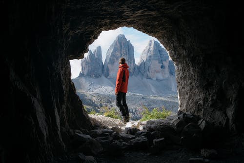 红色外套的人站立在三座山前面的洞的外部 · 免费素材图片