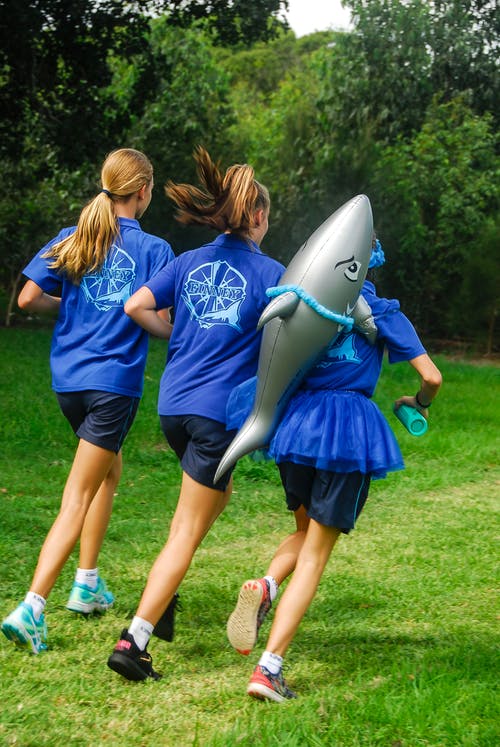 一个女孩背着鲨鱼气球和两个穿着蓝色制服的女孩跑 · 免费素材图片
