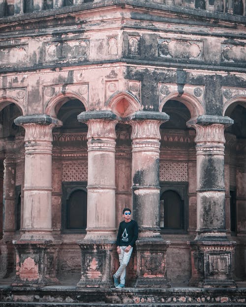 黑色长袖衬衫和白色牛仔牛仔裤的男人，白天斜倚在混凝土建筑上 · 免费素材图片