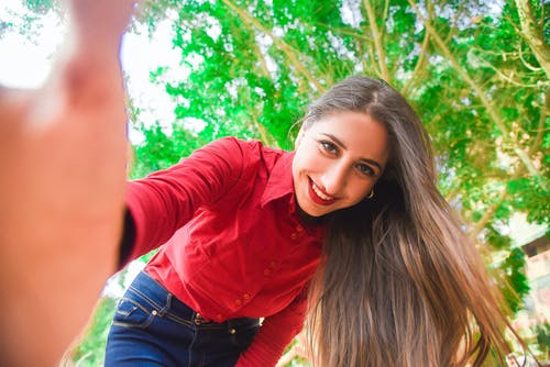 微笑的女士穿着红色衬衫和蓝色牛仔裤采取自拍照在绿叶树下 · 免费素材图片