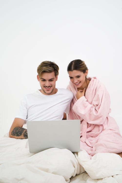 坐在白色的圆领衬衫的男人坐在床上旁边粉红色浴袍看着手提电脑的女人 · 免费素材图片