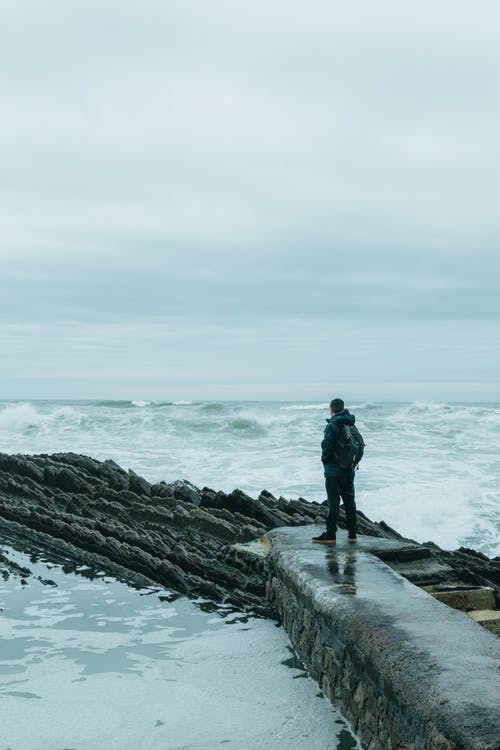 穿黑夹克的男人站在水体附近的岩层上 · 免费素材图片