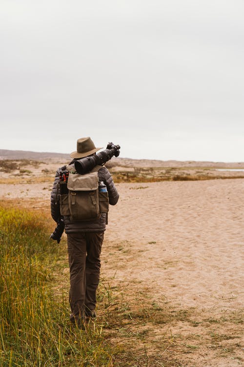 携带相机设备在沙地上行走的人的背影 · 免费素材图片