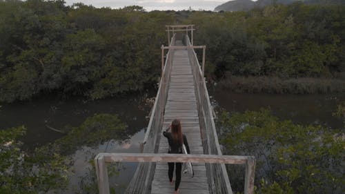 一名妇女背着冲浪板越过一座小木桥，通往树林的慢动作镜头 · 免费素材视频