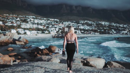 一个女人背着瑜伽垫，走到海边岩石上的背面 · 免费素材视频