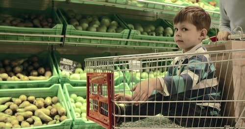 一个男孩被放在推车里，穿过杂货店的蔬菜区 · 免费素材视频